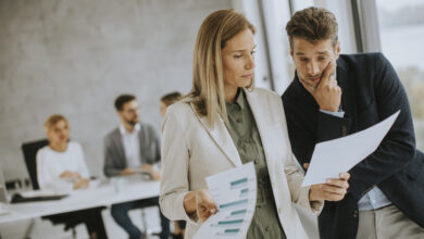 Mann und Frau diskutieren mit Papier in den Händen drinnen im Büro mit jungen Menschen arbeitet hinter ihnen.