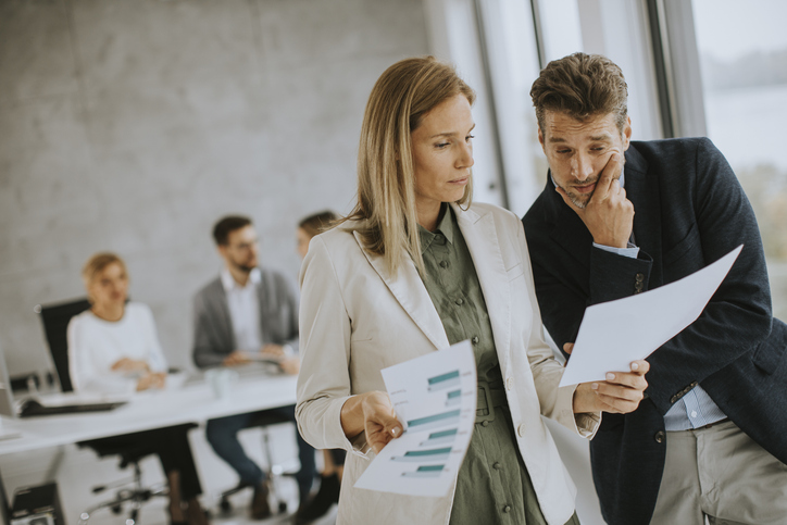 Mann und Frau diskutieren mit Papier in den Händen drinnen im Büro mit jungen Menschen arbeitet hinter ihnen.